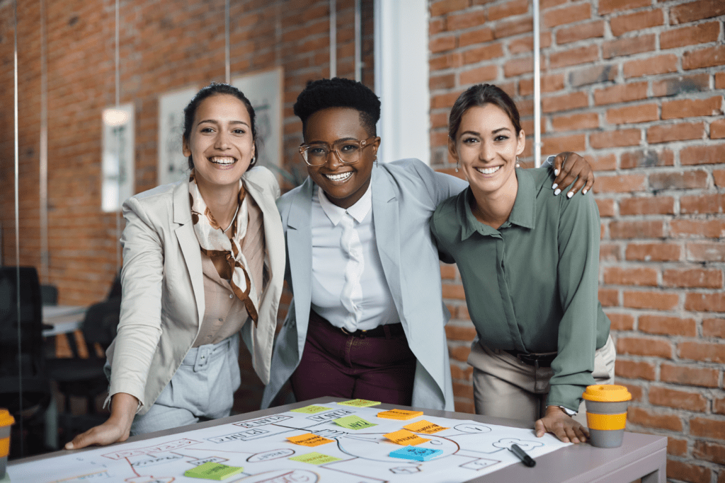 3 women of different ethnicity with their arms around each others shoulders, leaning on a table with a business mindmap