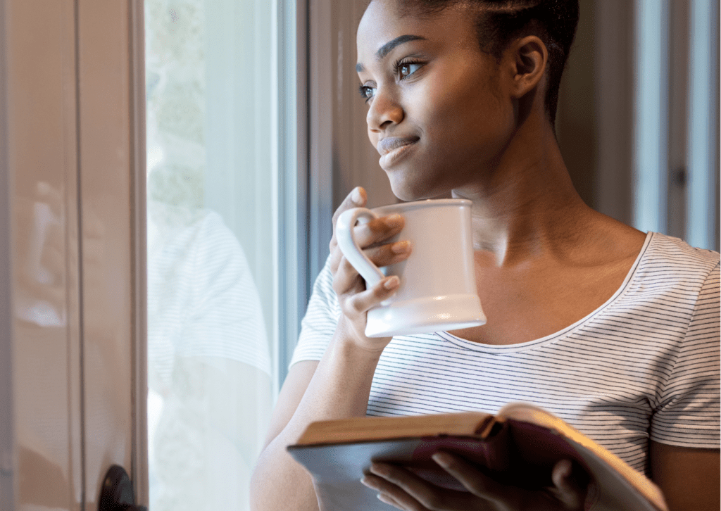 Black women gazing reflectively out of a window whilst holding a mug and a journal
