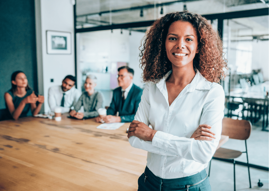 Black women dressed in a white shirt and trousers with arms folded. She is smiling and has shoulder length curly hair. She is standing in front of a meeting with 3 people in the background