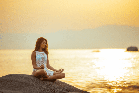 Lady sitting on a rock with legs crossed and hands resting on knees by water. Looking serene and connecting with her body